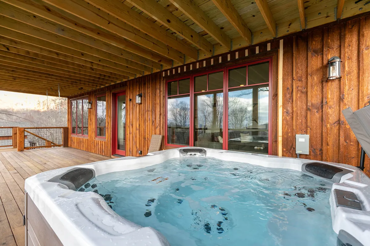 An outdoor scene featuring a bubbling hot tub on a wooden deck, set against large windows and surrounded by nature. The area looks serene yet sociable—perfect for soaking in mountain views or enjoying company in the fresh air.