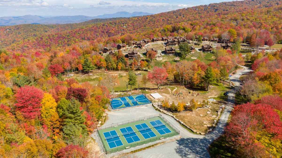 A vibrant autumn exterior image showcasing tennis courts, colorful fall foliage, cabins, and mountain views. The scene is lively and energetic, hinting at a range of outdoor recreational opportunities and natural beauty within the resort.
