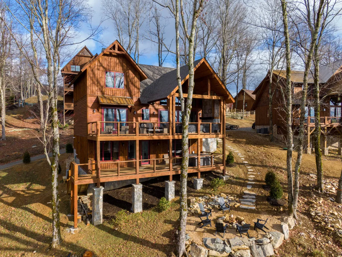 The image showcases the exterior of Firesky Lodge, featuring a spacious wooden deck and large windows that invite the mountain landscape inside. A stone patio sits nestled among tall trees, creating a serene backdrop for outdoor gatherings. The sunset casts a warm glow over the balcony, and a firepit offers a perfect spot for stargazing or sharing stories under the night sky.