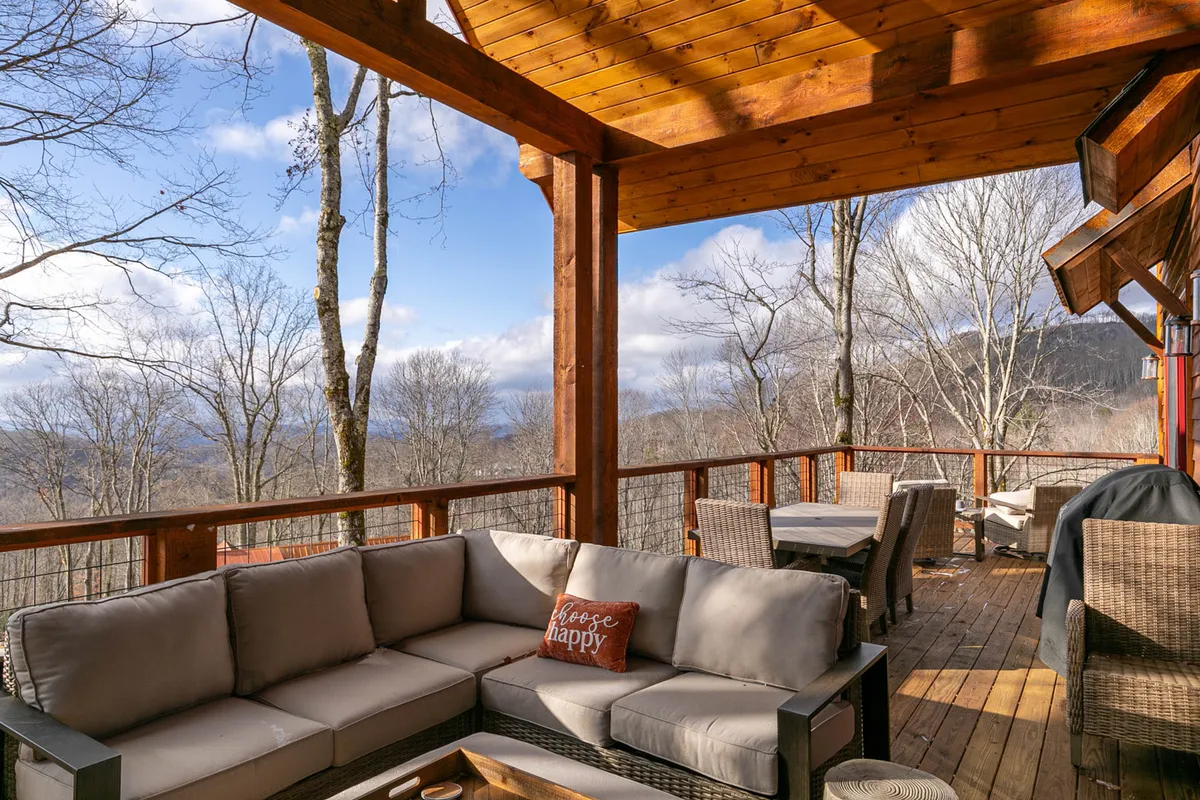 A cozy, comfortable outdoor seating area under wooden beams, overlooking a picturesque view of trees and mountains. The arrangement suggests moments of relaxation, enjoying coffee at sunrise, hosting small gatherings, or simply soaking in the serenity.