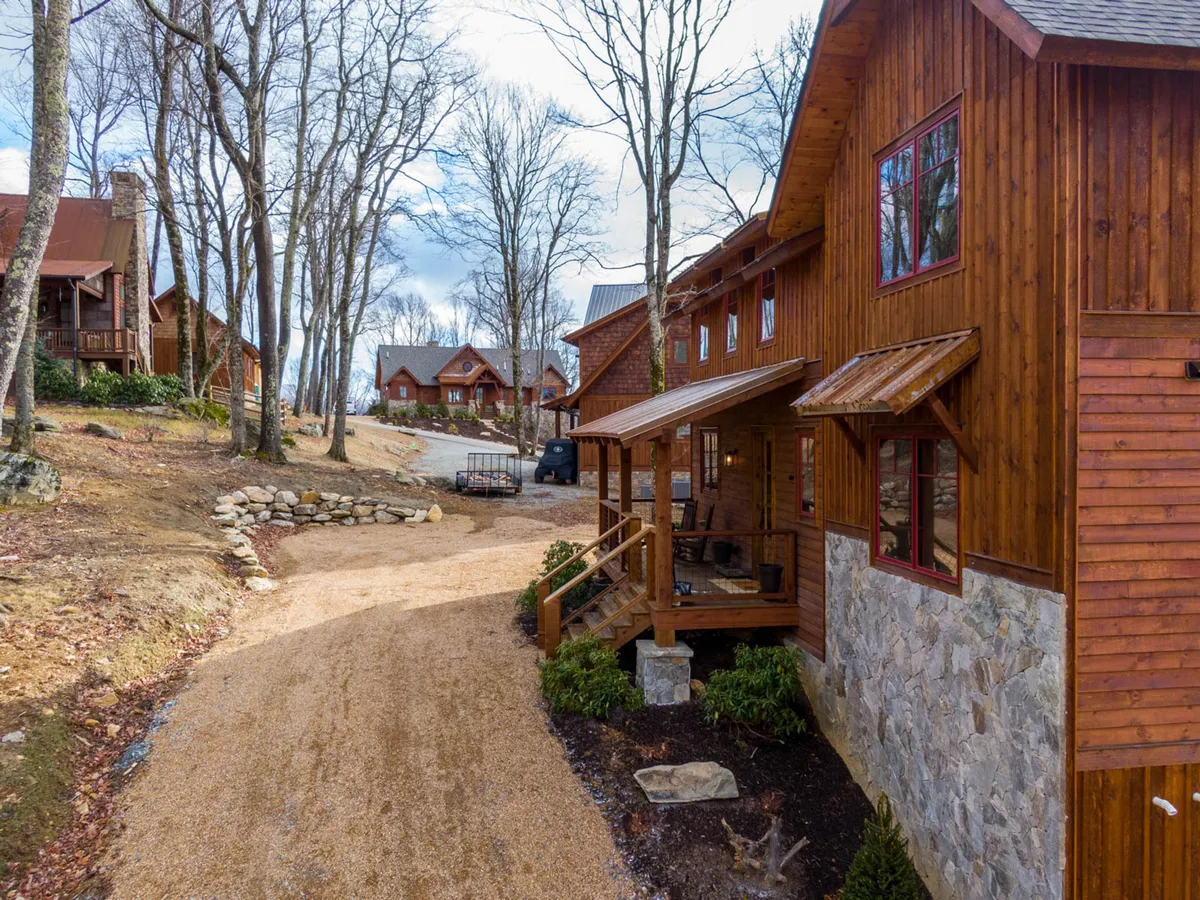 This image captures the wooden exterior of the house, a stone pathway leading to the porch, and a view of the surrounding trees and nearby cabins. The scene is tranquil and suggests easy access to nature walks and community spaces.