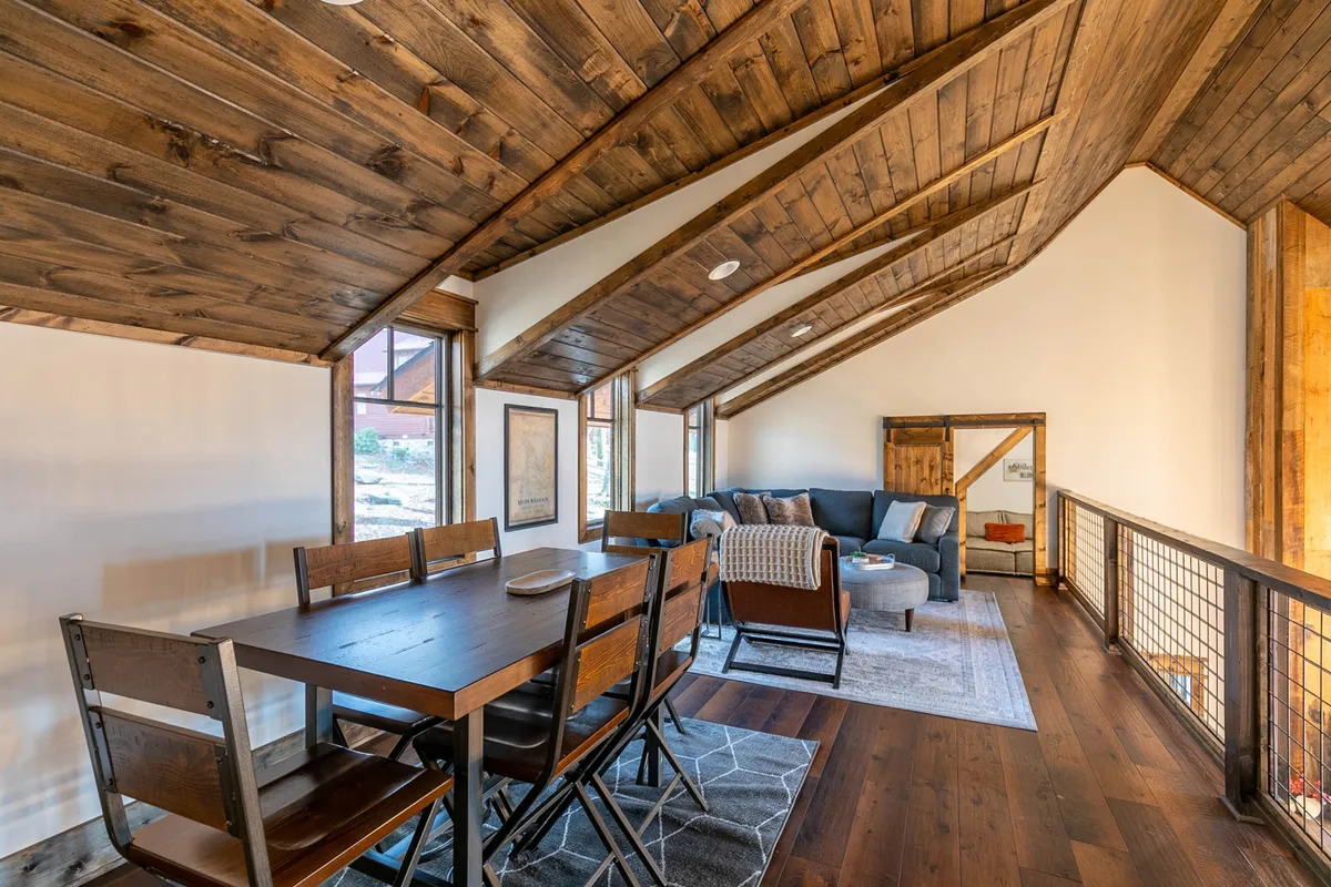 A spacious great room with rustic wooden beams overhead, plush seating including a sofa arranged for conversation, a large dining table ready for family dinners, and a dramatic wall of windows framing the Blue Ridge mountain views. Warm, ambient lighting and welcoming decor set an inviting, communal scene.