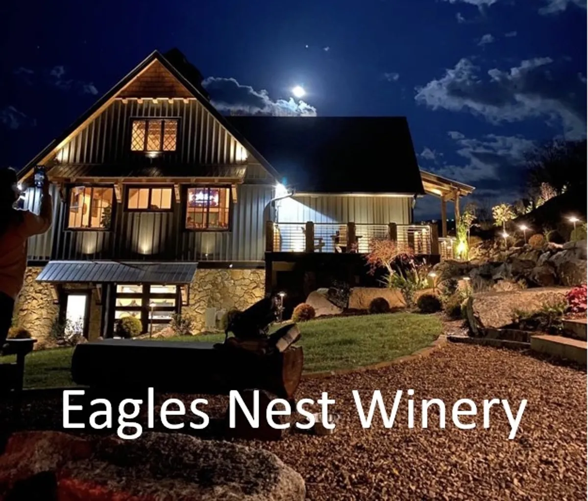 An exterior evening shot featuring the illuminated building of the lodge, a glowing moon overhead, and a beautifully landscaped garden in the foreground. Soft lighting suggests an inviting, romantic ambiance, and the setting hints at relaxed outdoor gatherings or wine tastings under the stars.