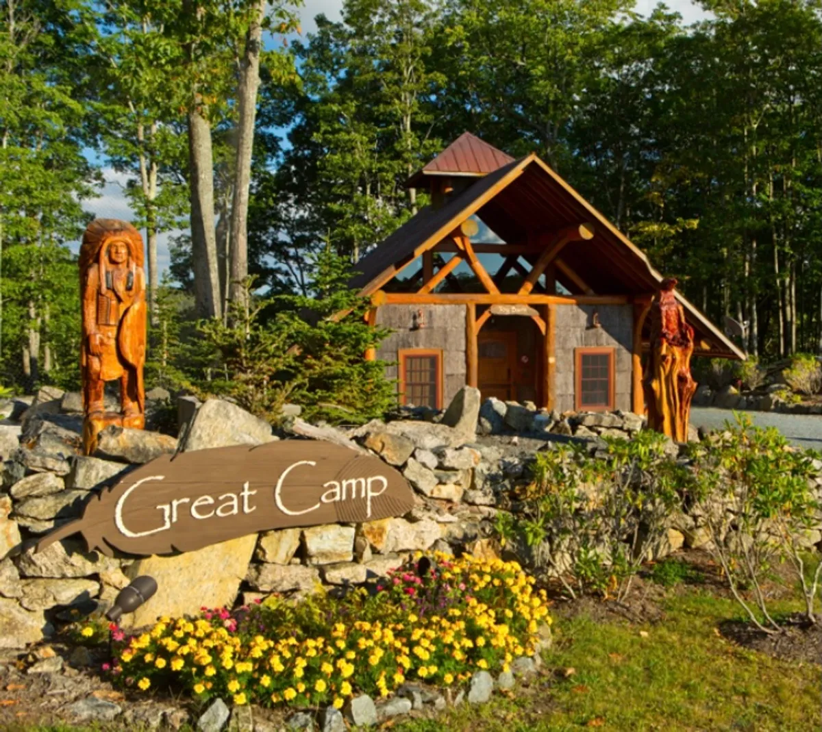 The image displays the main cabin with charming wooden sculptures, a stone wall, and colorful flower beds in the foreground. The setting highlights an inviting and rustic outdoor space ideal for gathering and play.