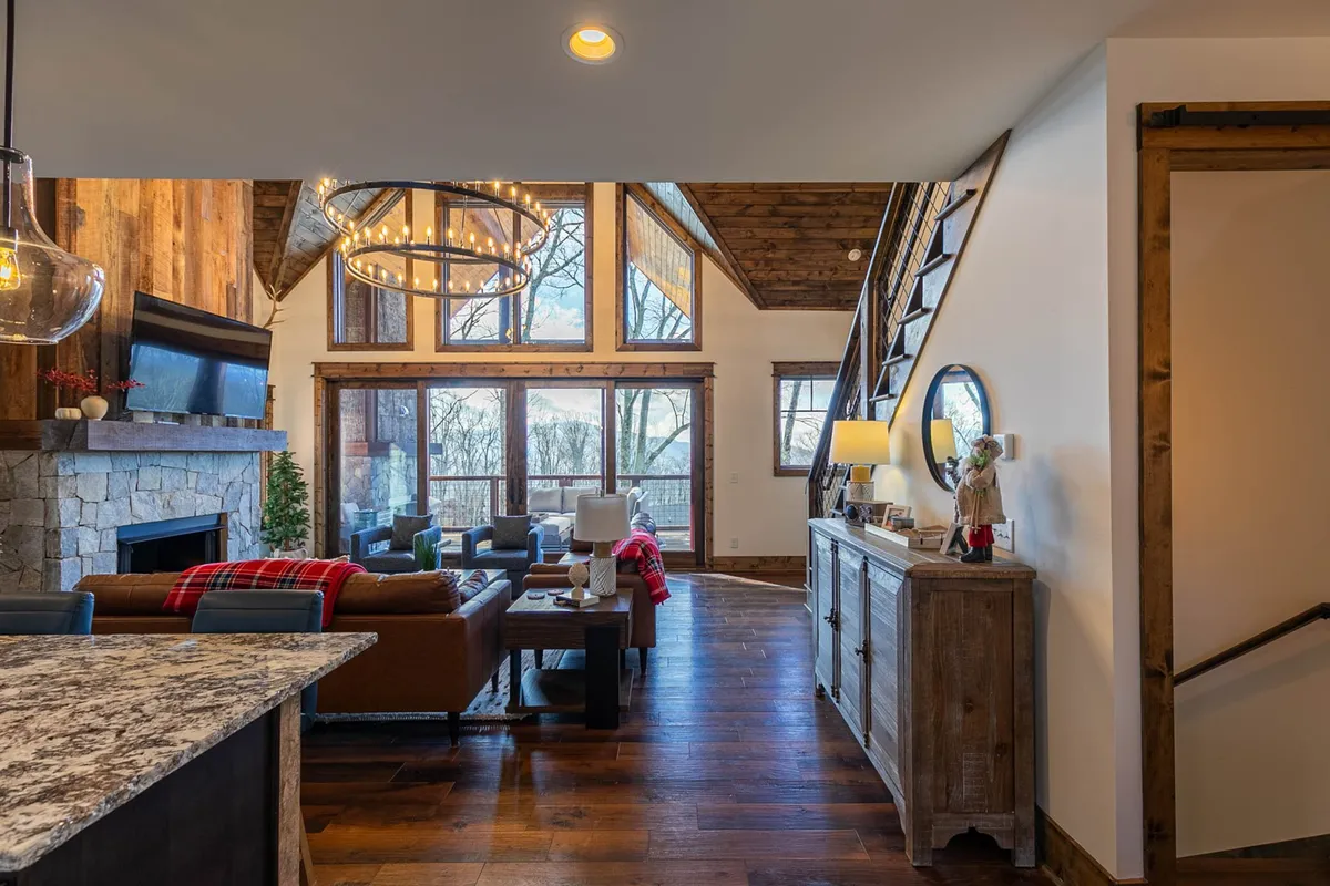 A bright living room with expansive windows offering sweeping mountain views, a rustic stone fireplace, and comfortable seating for guests. The space features warm wooden beams and a relaxed, inviting environment that emphasizes a connection to the outdoors and the surrounding natural beauty.