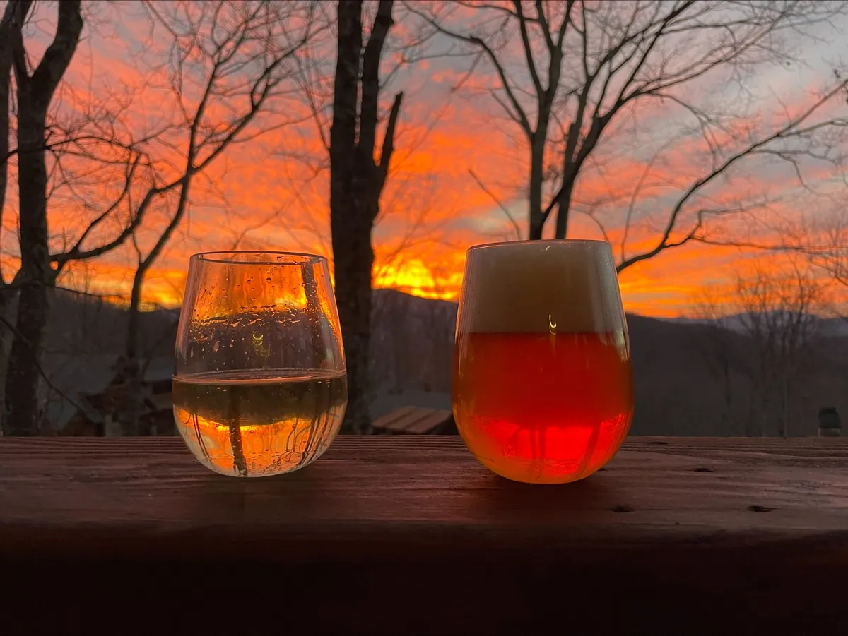 A pair of glasses—one with clear liquid and one with beer—sit on an outdoor table with a breathtaking sunset sky and tree silhouettes in the background. The mood is relaxed and sociable, evoking moments of connection among friends and family in a beautiful mountain setting.