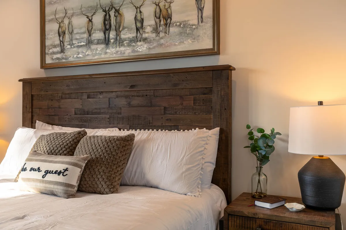 A cozy bedroom with a wooden headboard, colorful decorative pillows, animal artwork, a side table with a lamp, and a small plant. The bed looks inviting and is set against a backdrop of soft natural light from a nearby window.