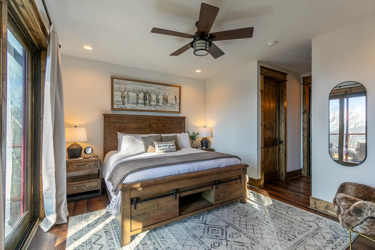 A tranquil bedroom with a large bed, wooden nightstands, and soft artwork above the headboard. Light streams in from the window, illuminating cozy linens, and there's a mirrored accent that adds depth to the room. The overall mood is calm and restful.