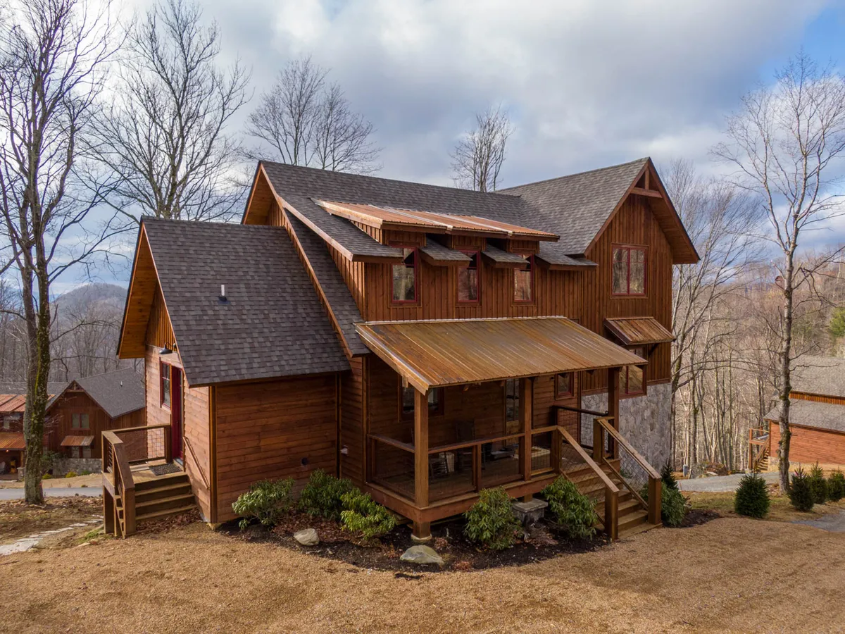 The image showcases the lodge's wooden exterior with a spacious porch, surrounded by tall trees and natural stone accents. The setting evokes a cozy, welcoming atmosphere ideal for relaxing outdoors with pets or gathering after a day of adventure.
