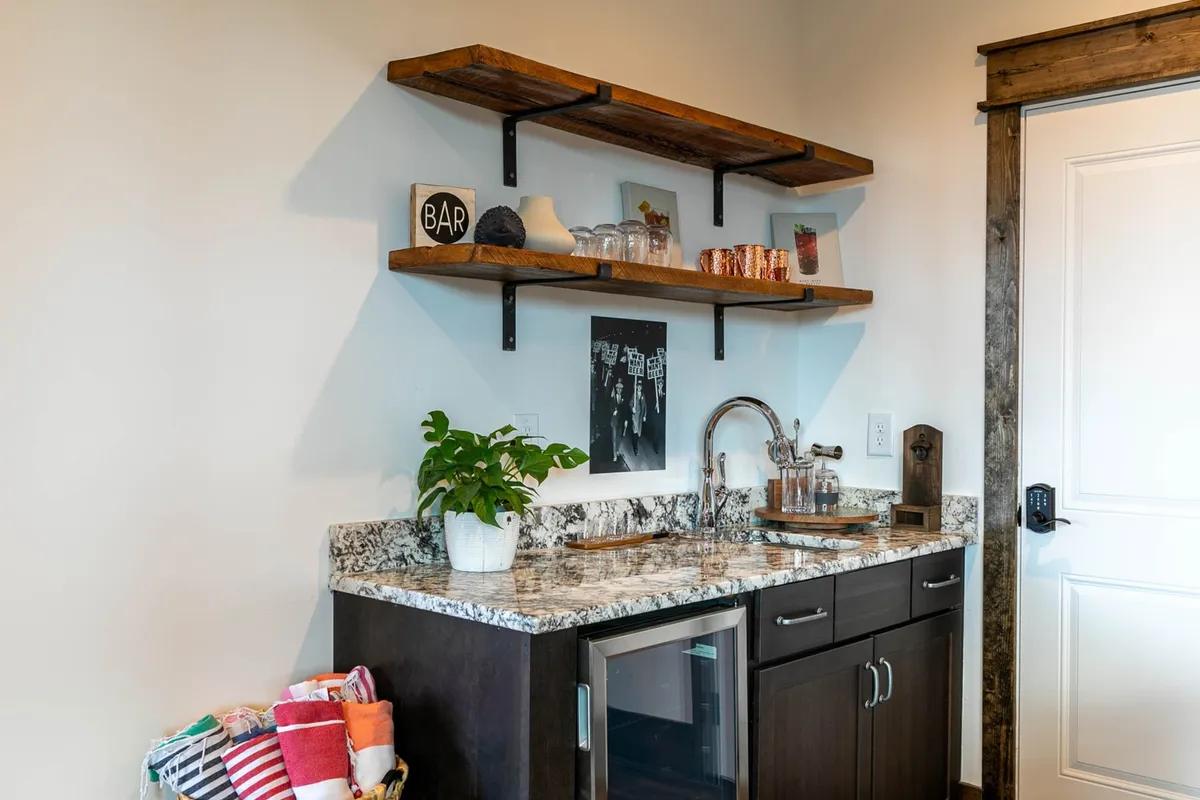 A welcoming wet bar area with granite countertops, wooden shelves, a bar sign, potted plant, and an array of glassware. The setup suggests a social atmosphere ideal for mixing drinks and sharing stories during gatherings.
